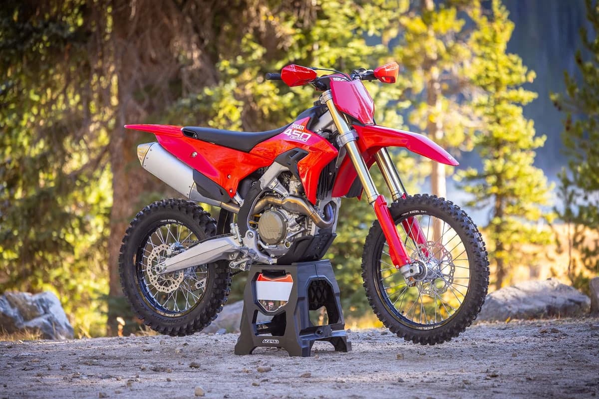 2026 Ducati Desmo450 EDX in red and black, left-side studio profile on a wooden stand showing gold forks, spoke wheels and a silver muffler