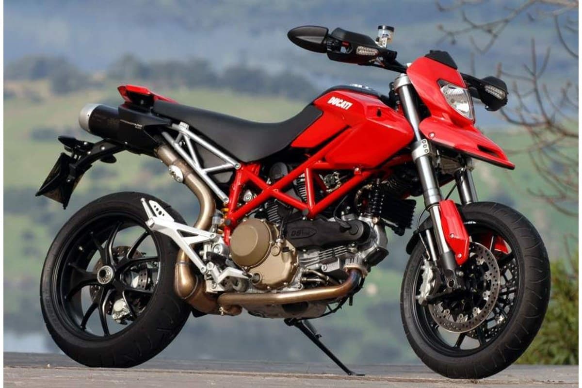 2012 Ducati Hypermotard 1100 in red, right-side outdoor profile showing the trellis frame, high-mounted exhaust and L-twin engine