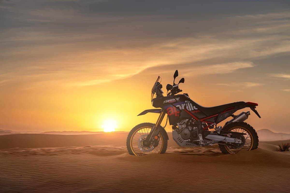 2027 Aprilia Tuareg 660 Rally parked on a desert dune at sunset, silhouetted against a glowing orange sky with the gold forks and knobby tires catching the light