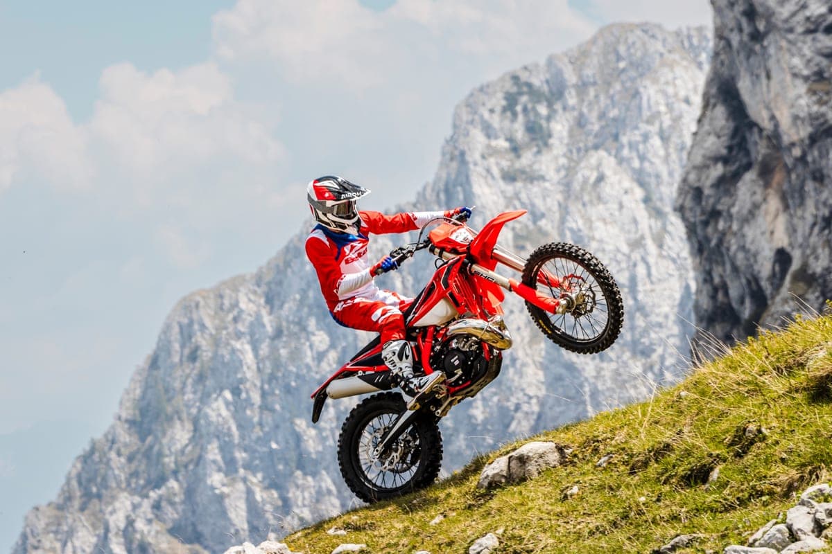 Beta 200 RR X-Pro popping a wheelie on a rocky mountainside, rider in red-and-white gear against jagged grey cliffs and green scrub