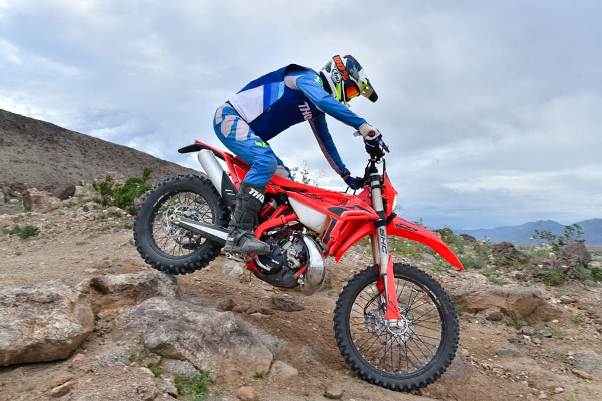 Beta 200 RR X-Pro cresting a rocky hillside climb, rider in blue gear leaning forward with cloudy desert mountains in the background