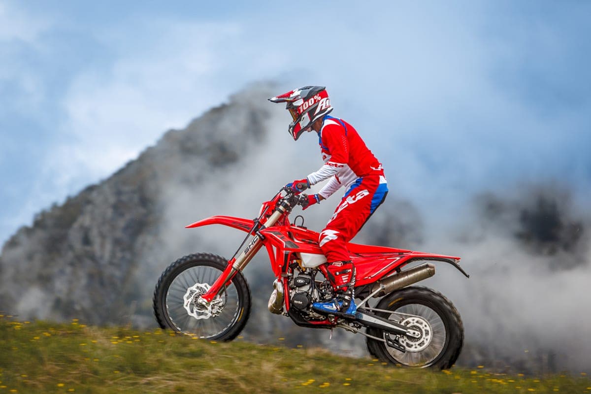 Rider in red and blue Beta gear popping the front wheel up a grassy slope on a 250 RR X-Pro, smoke drifting behind against a mountain backdrop