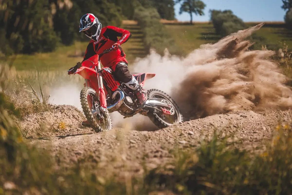 Rider in matching red and black gear powering a Beta 250 RX out of a dirt berm, kicking up a huge roost of sandy soil