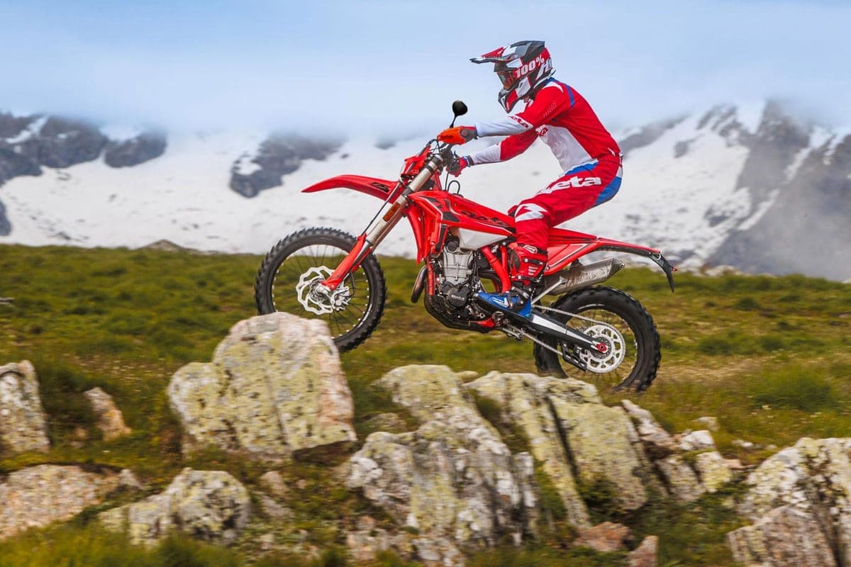 Rider in red and white Beta gear catching air over rocky terrain on a Beta 350 RS, with snow-capped mountains behind