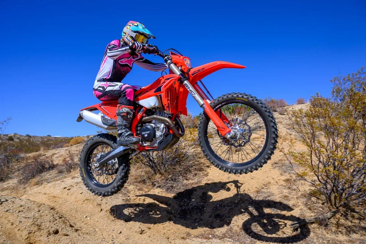 Rider in pink and white gear launching a red Beta 390 RR X-Pro off a desert jump, front wheel high and dirt kicking off the rear knobby tire