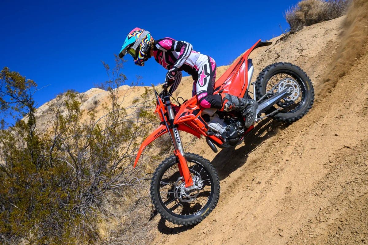 Rider in pink and white gear leaning a red Beta 390 RR X-Pro down a steep sandy hillside, rear tire carving a dust trail under a clear blue sky