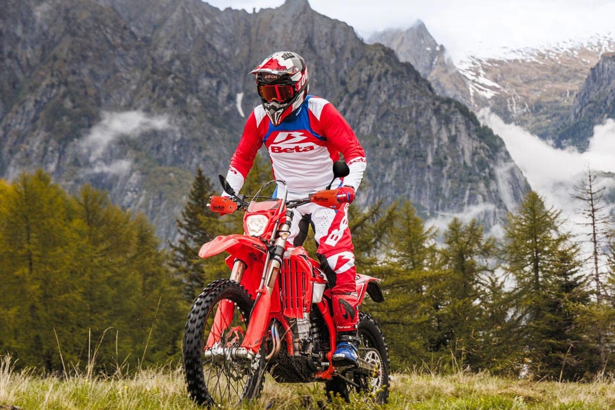 Rider in red and white Beta gear astride a red 390 RS on a grassy alpine slope with pine forest and snow-capped peaks behind