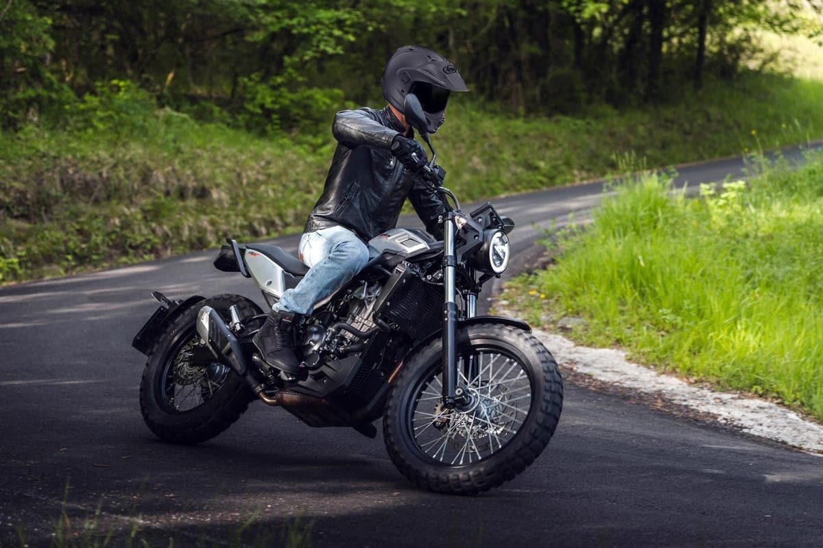 Rider in a black leather jacket and jeans riding a Beta Alp X down a tree-lined country road, front wheel lifted slightly off the pavement