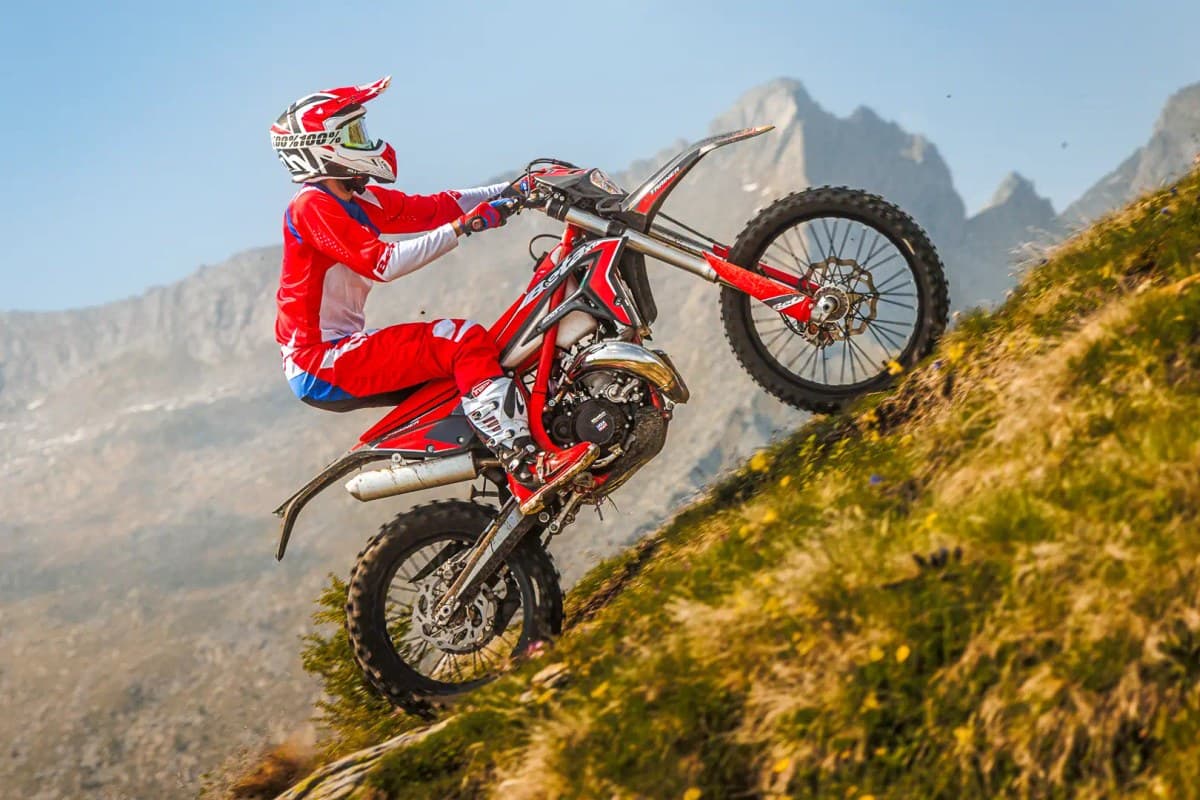 Rider in red and white gear powering the Beta Xtrainer 300 up a steep grassy mountainside, front wheel lofted with rocky peaks behind