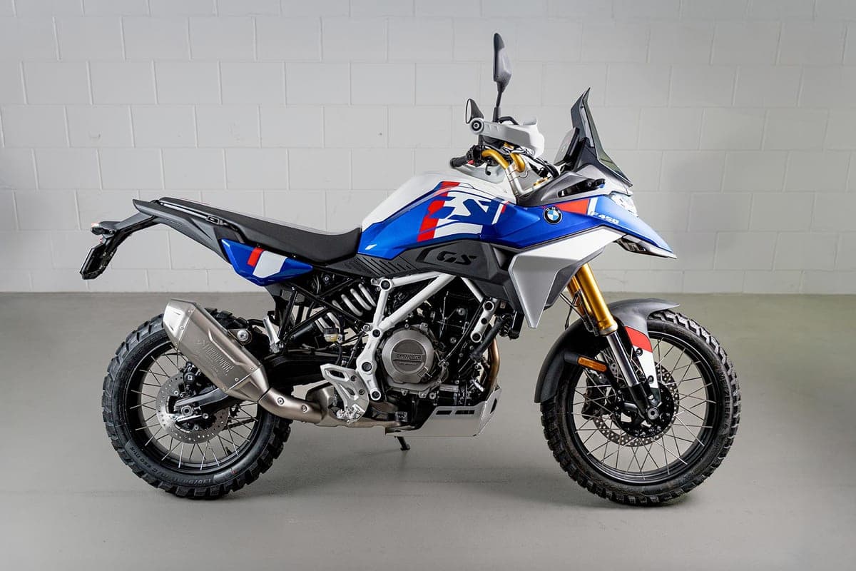 2027 BMW F 450 GS in white, blue and red livery, right-side studio profile against a brick wall highlighting the Akrapovic-style exhaust and knobby tires