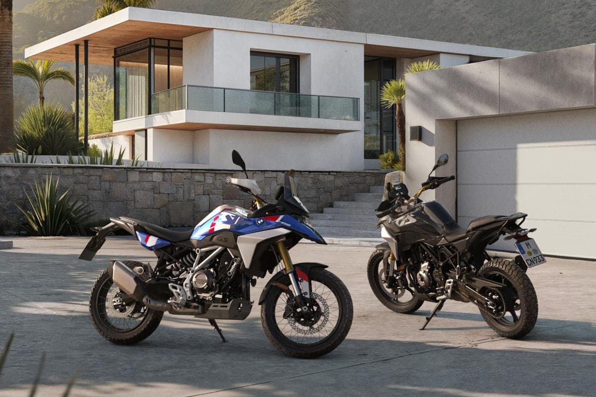 2027 BMW F 450 GS and a grey sibling model parked rear three-quarter outside a modern stone-and-glass house, showing the tall tail section and license plate mount