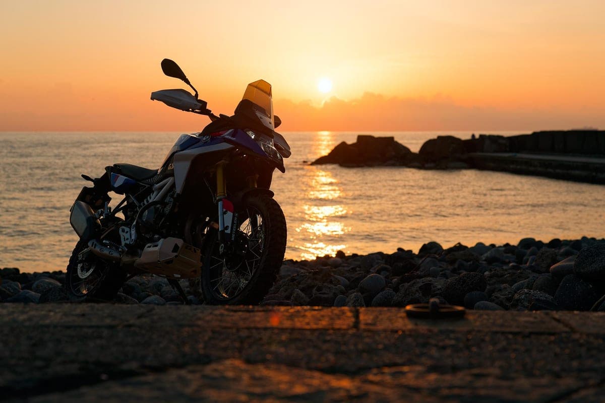 BMW F 450 GS silhouetted front three-quarter on a rocky shoreline at sunset, windscreen and headlight cowl backlit by the glowing horizon