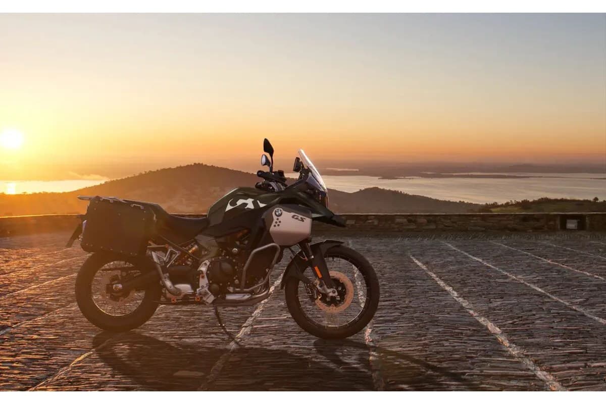 BMW F 900 GS Adventure in camo green with white GS panel, parked on a stone overlook at sunset with a lake and hills in the background