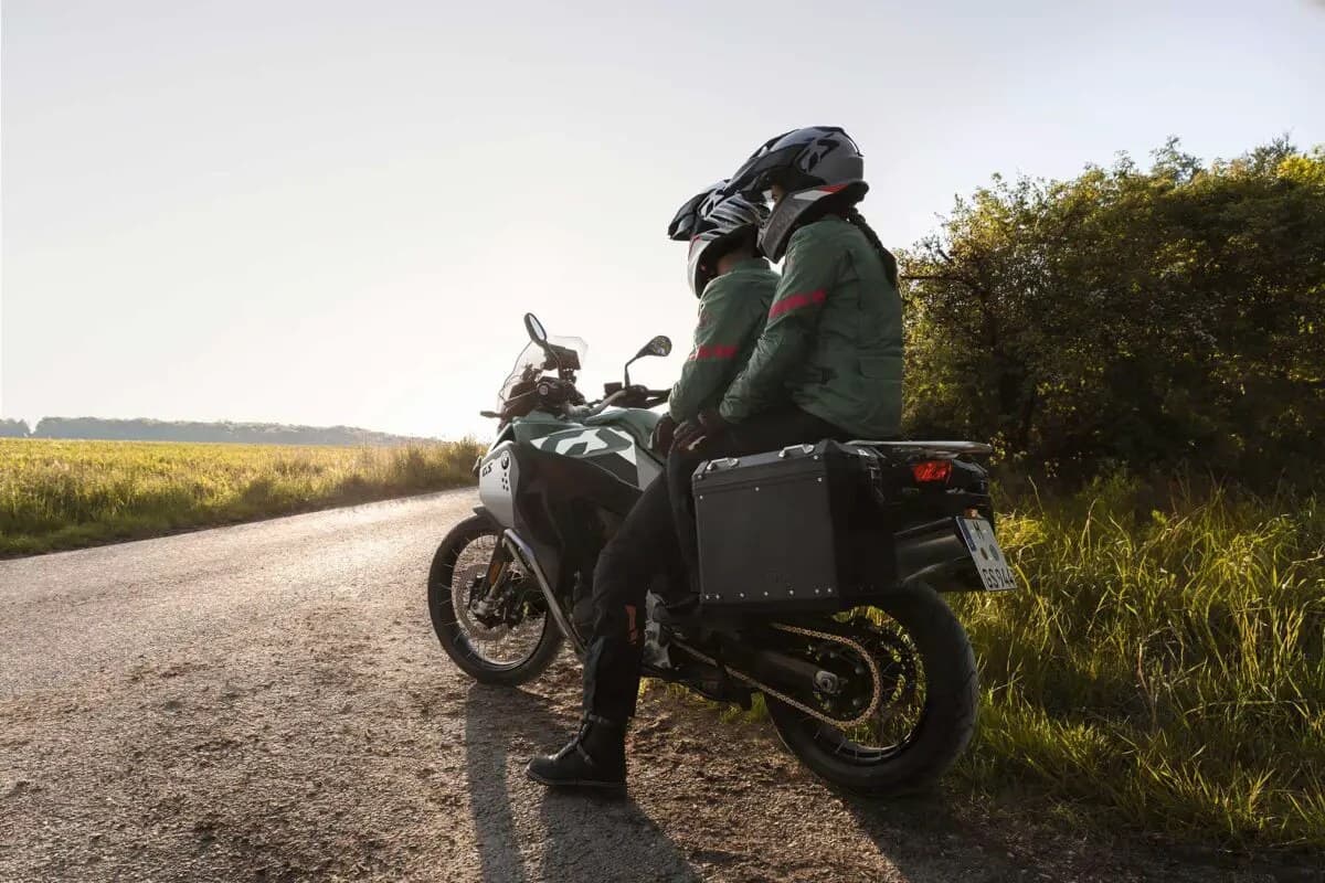 Two riders in green touring gear seated on a BMW F 900 GS Adventure with side cases, pausing on a gravel road backlit by low sun