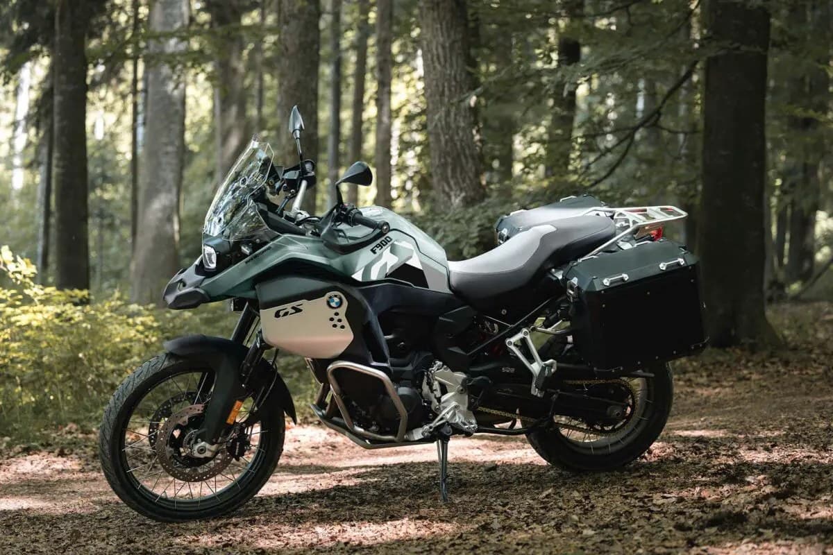 BMW F 900 GS Adventure in sage green with silver GS badge, front three-quarter view on a forest trail showing the crash bars and panniers