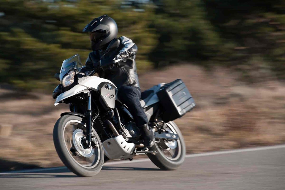 Rider in black leather jacket and full-face helmet riding a white BMW G650GS with side pannier down a sunlit country road