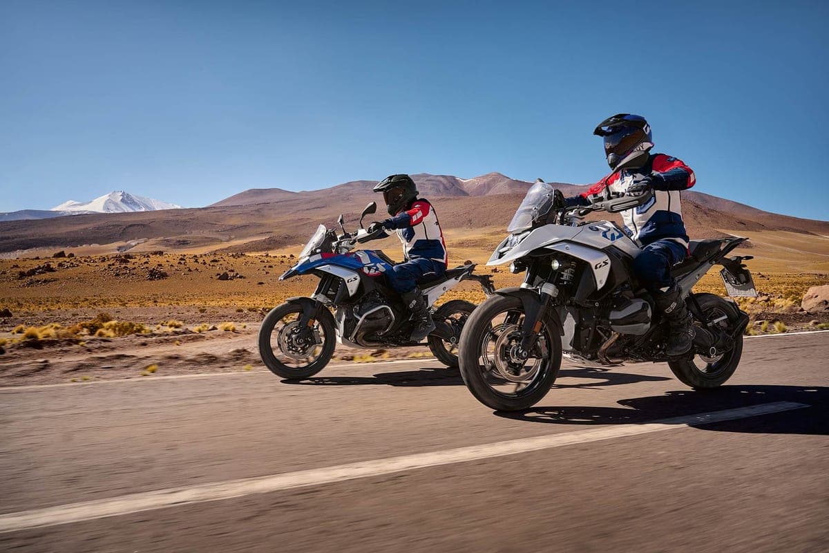 Two riders in GS-branded gear riding blue and white BMW R 1300 GS motorcycles along a desert highway with snow-capped mountains behind them
