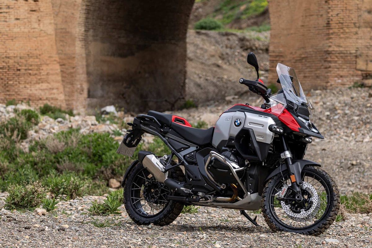 BMW R 1300 GS Adventure parked on gravel beneath a stone bridge arch, front three-quarter view highlighting the knobby tires and skid plate off-road