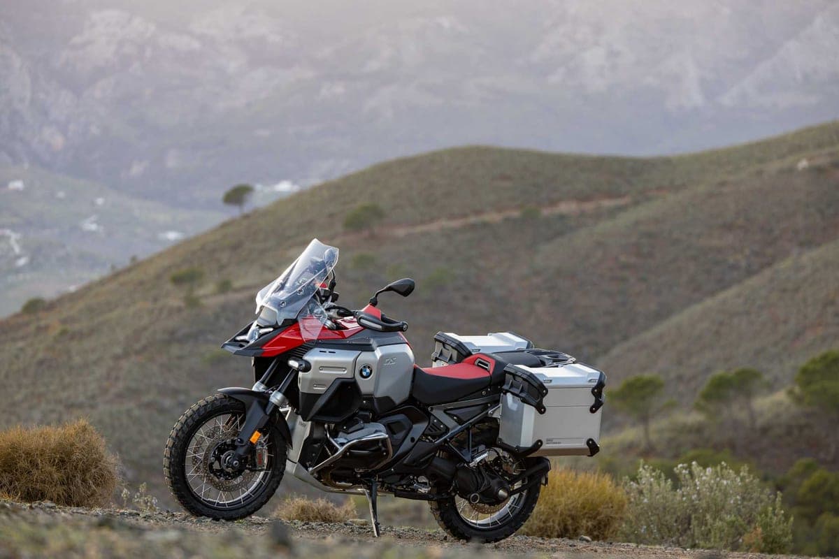 BMW R 1300 GS Adventure with aluminum panniers fitted, rear three-quarter view on a hilltop overlooking layered mountain ridgelines at dusk
