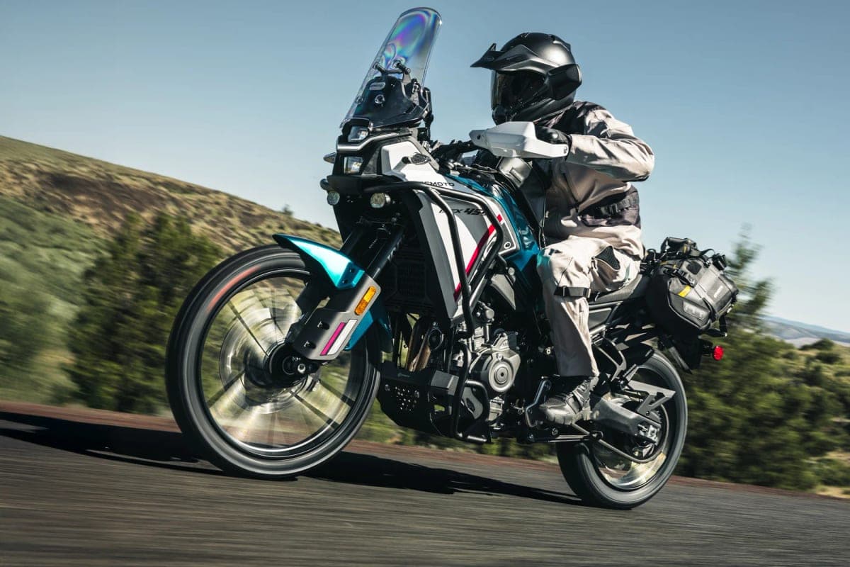 Rider in tan riding gear on a blue and white CFMoto Ibex 450 popping a wheelie on pavement, soft-luggage panniers strapped to the tail