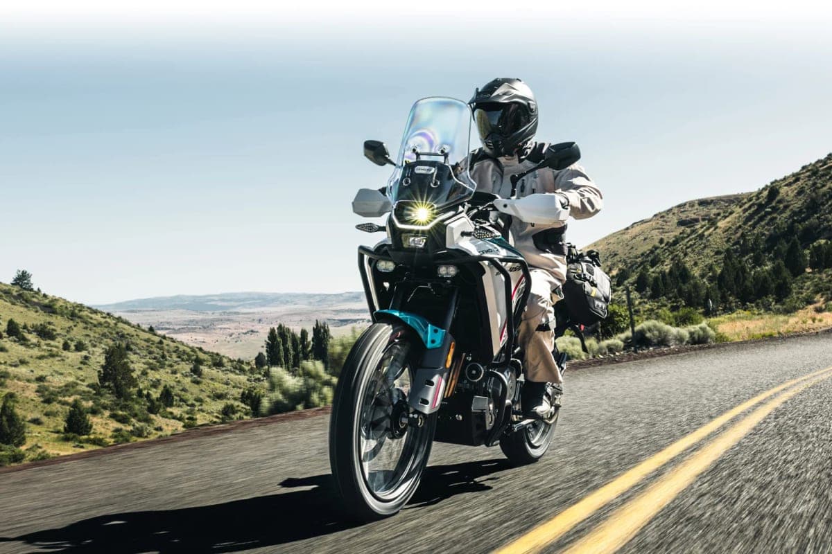 Rider in tan gear on a CFMoto Ibex 450 riding toward camera on a mountain road, LED headlight glowing beneath the tall windscreen