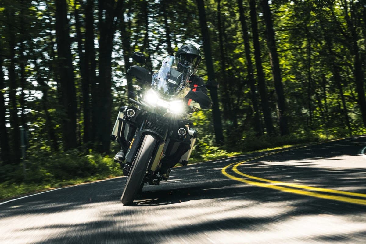 CFMoto Ibex 800 E ridden head-on toward the camera at dusk with LED headlights blazing, rider tucked behind the windscreen on a shaded tree-lined curve