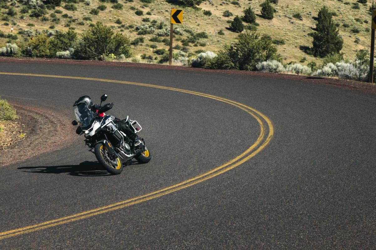White and gold CFMoto Ibex 800 E rear three-quarter action shot, rider cornering hard on a desert switchback with a yellow chevron road sign behind