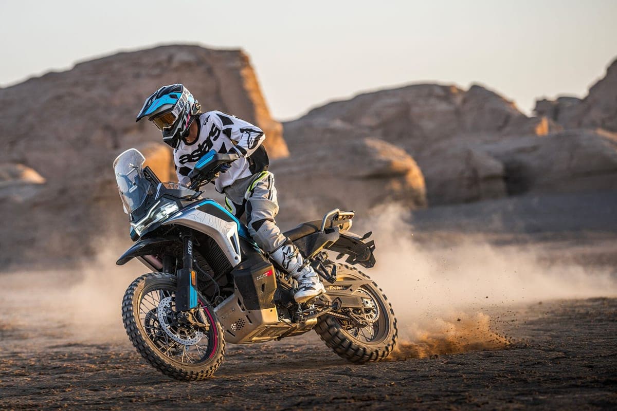 Rider in white and black gear powersliding a CFMoto Ibex 950 through a desert canyon, kicking up a large plume of dust at sunset