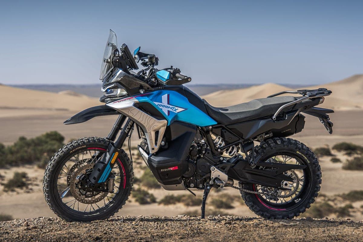 CFMoto Ibex 950 parked on a rocky overlook above sand dunes, side-on profile highlighting the tall windscreen, Brembo front brake and knobby tires