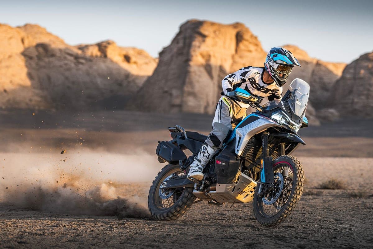 Rider in Alpinestars gear leaning a blue-and-white CFMoto Ibex 950 hard through a dusty desert turn, front wheel kicking up rocks and dirt