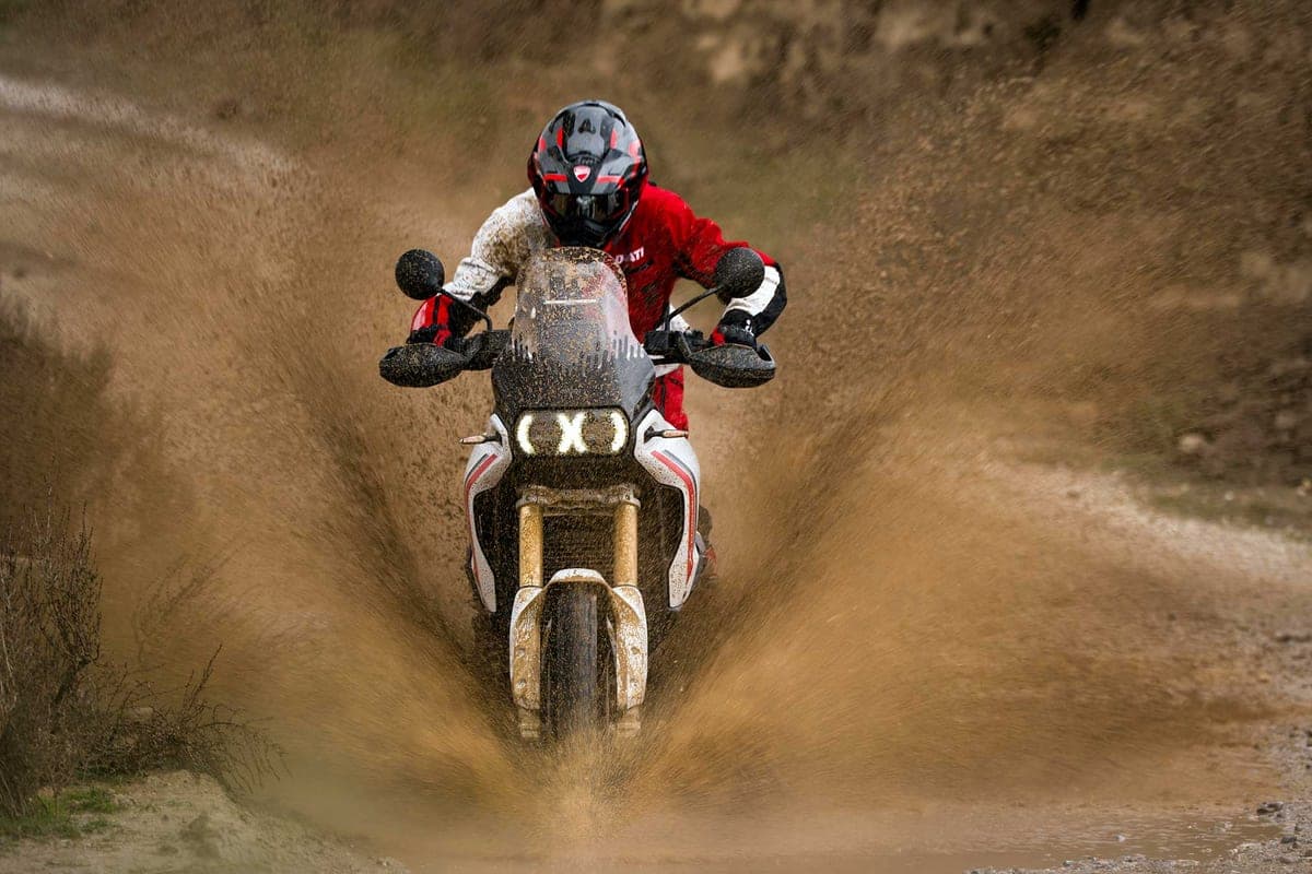 Rider in white and red Ducati gear powering the DesertX V2 through a muddy puddle, splashing water past the round LED headlight and beak fender