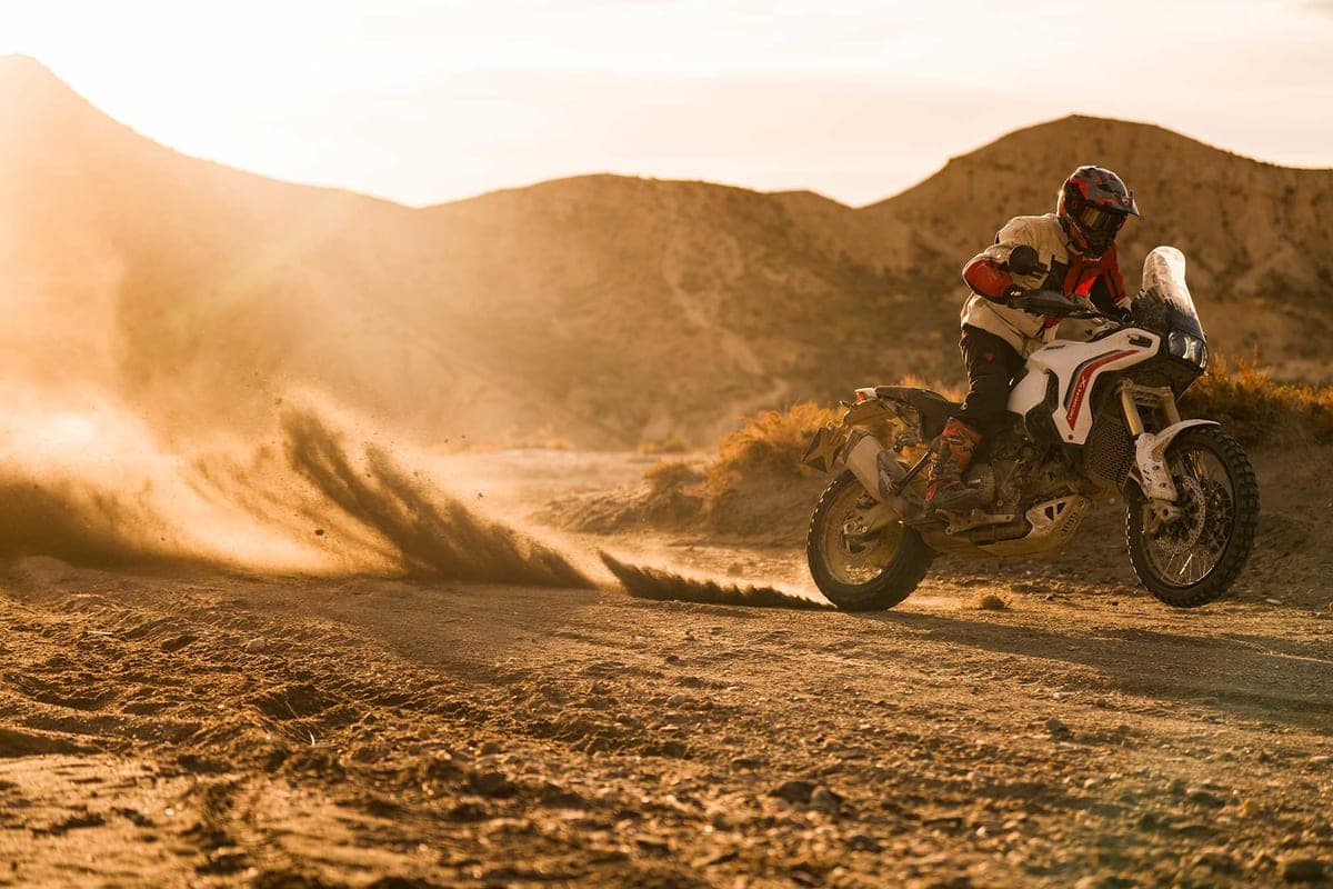 Rider on a white Ducati DesertX V2 kicking up a dust plume across a desert trail at golden hour, mountains silhouetted in the background