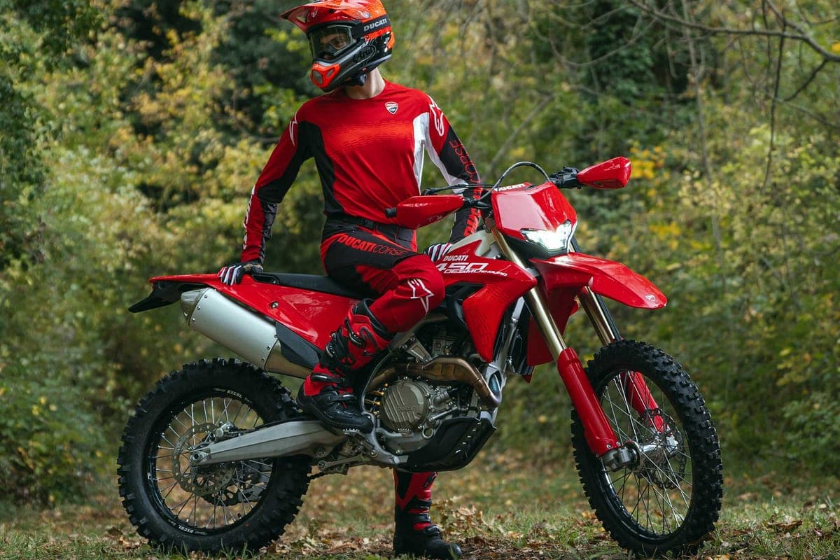 Rider in red and black Ducati Corse gear with an orange helmet standing over a red Desmo450 EDS in a wooded clearing, headlight lit