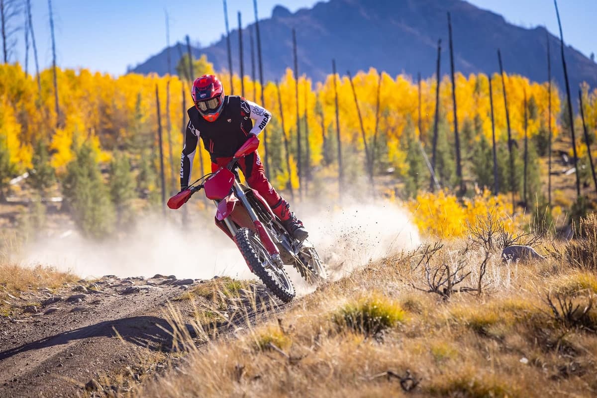 Rider in black and red Ducati gear railing the Desmo450 EDX around a dusty berm past golden autumn foliage and burnt tree trunks