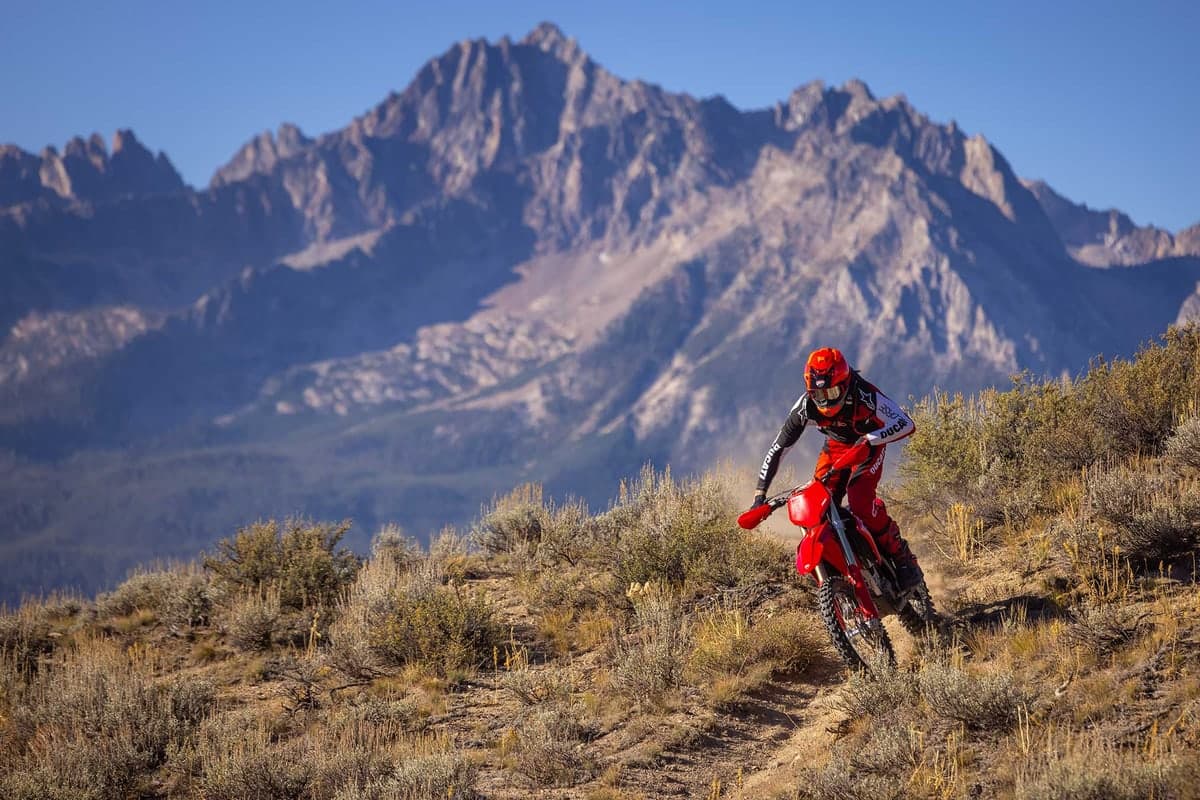 Ducati Desmo450 EDX rider leaning into a turn on a scrubby mountainside trail with jagged gray peaks filling the background