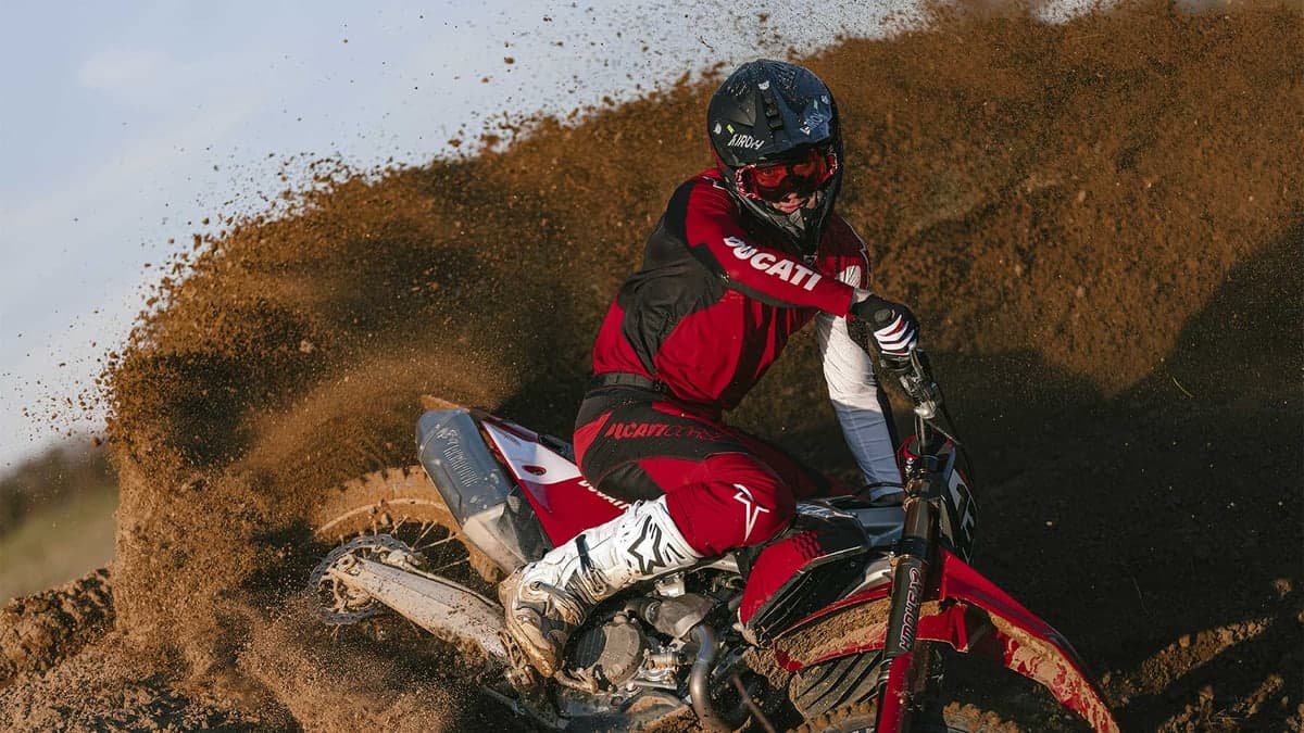 Ducati Desmo450 MX Factory leaning into a berm, rider in Ducati race gear and Airoh helmet carving through a wall of thrown dirt