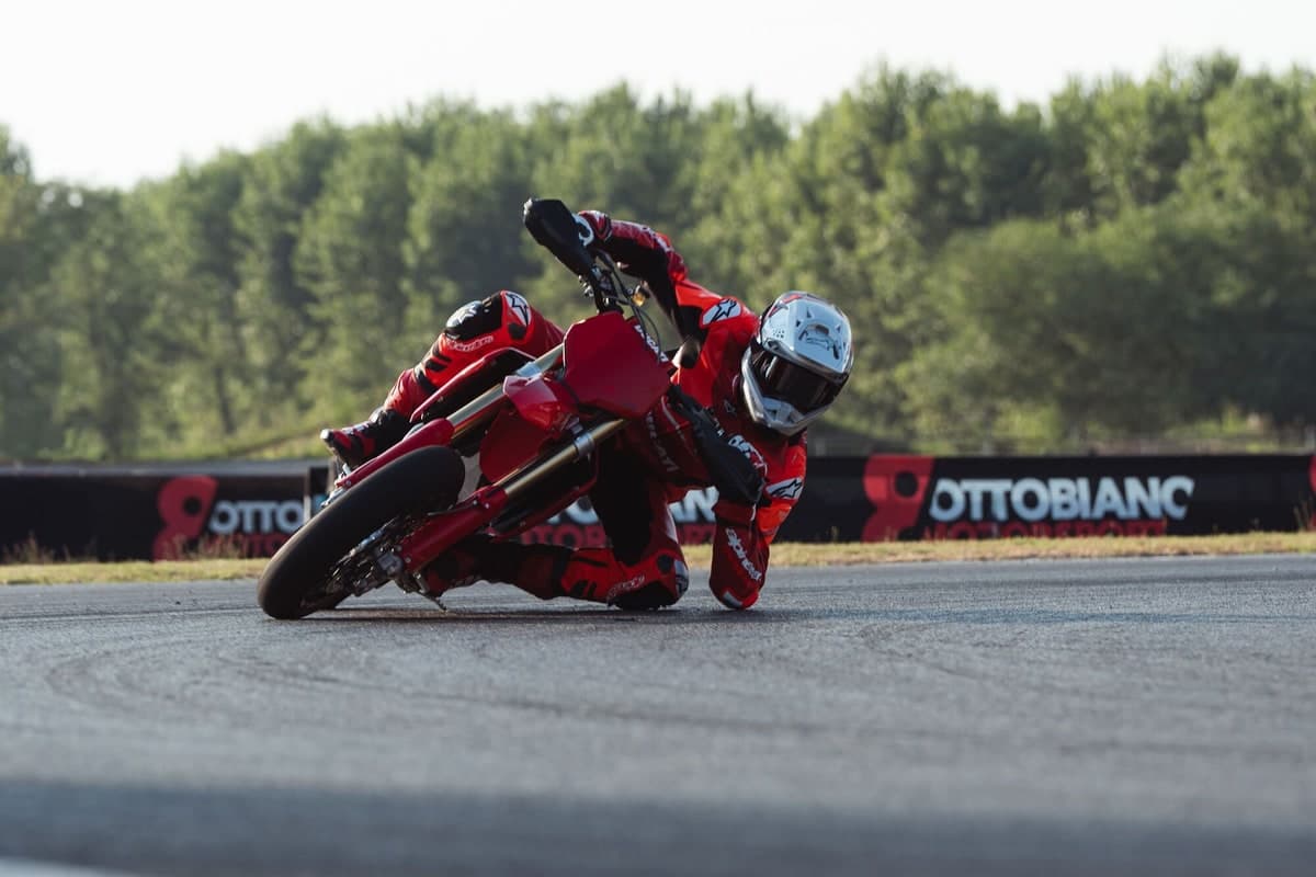 Rider in red Ducati leathers dragging a knee on the Desmo450 SM through a paved track corner, with a trackside banner behind