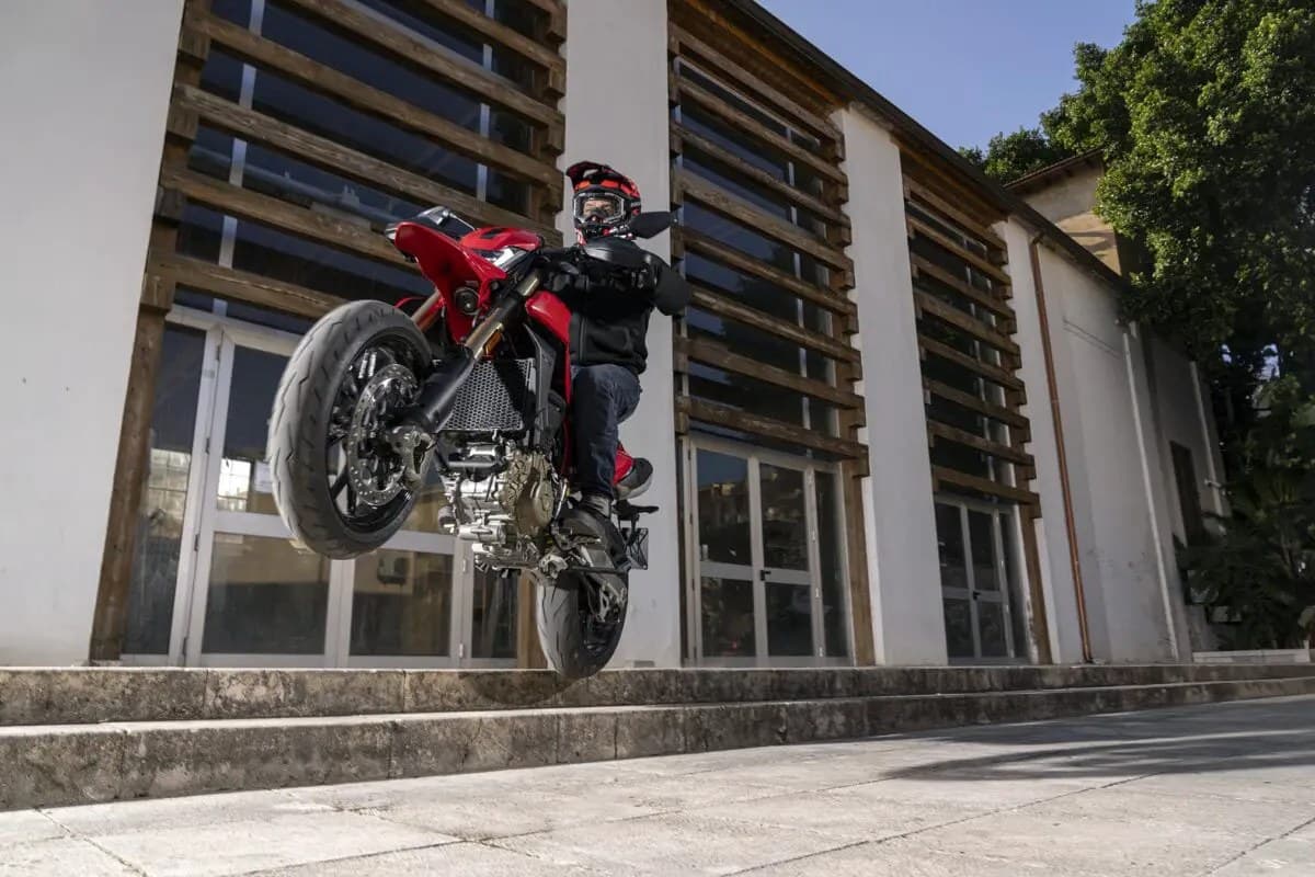 Rider in black gear on a red Ducati Hypermotard 698 Mono popping a wheelie past a slatted-wood building facade on a sunny street