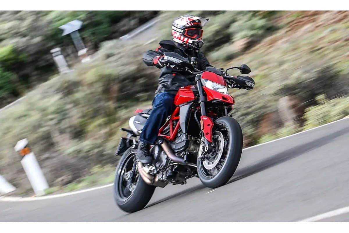 Rider in black leathers and a white helmet leaning the red Ducati Hypermotard 950 hard into a mountain road curve, front wheel lofted slightly