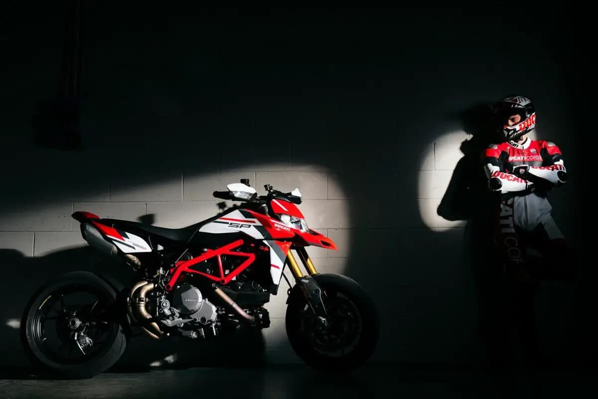 Ducati Hypermotard 950 SP parked in a dramatic spotlit garage beside a rider in Ducati Corse leathers and a full-face helmet, front three-quarter view