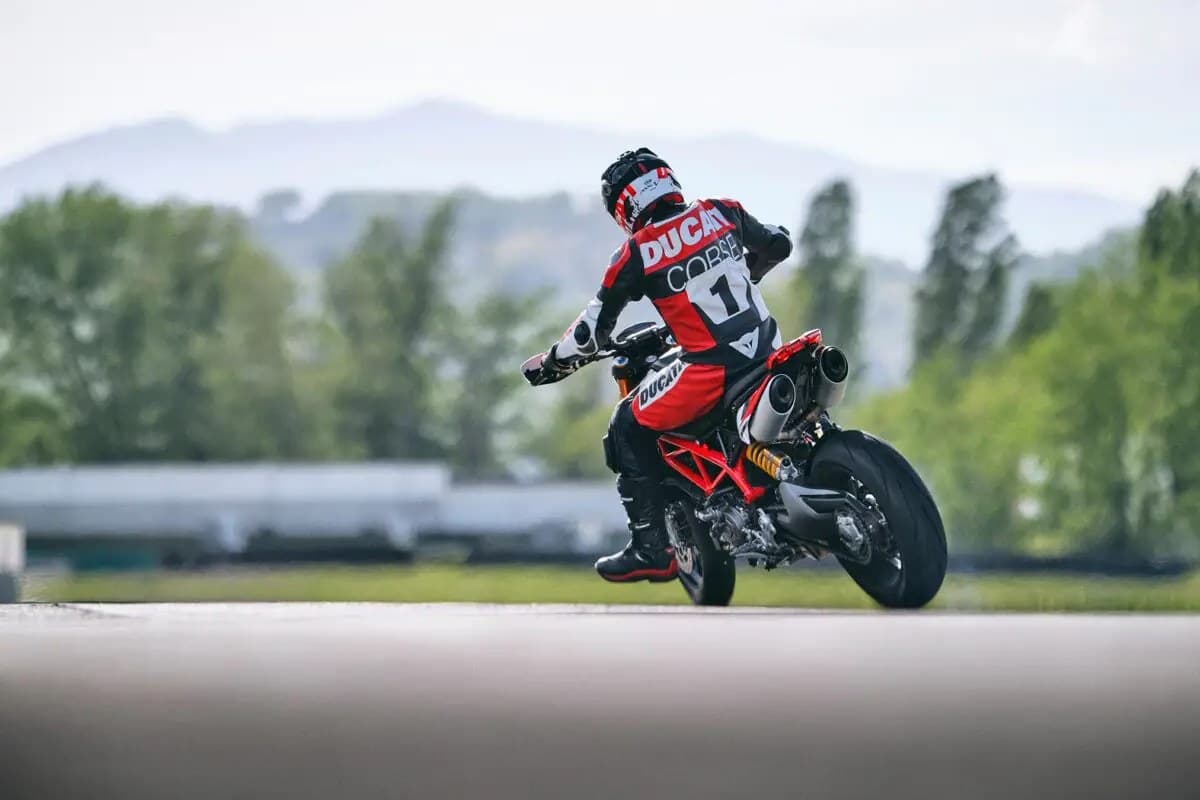 Rider in Ducati Corse race gear popping a wheelie on a red and white Hypermotard 950 SP down a track, trees and mountains blurred behind