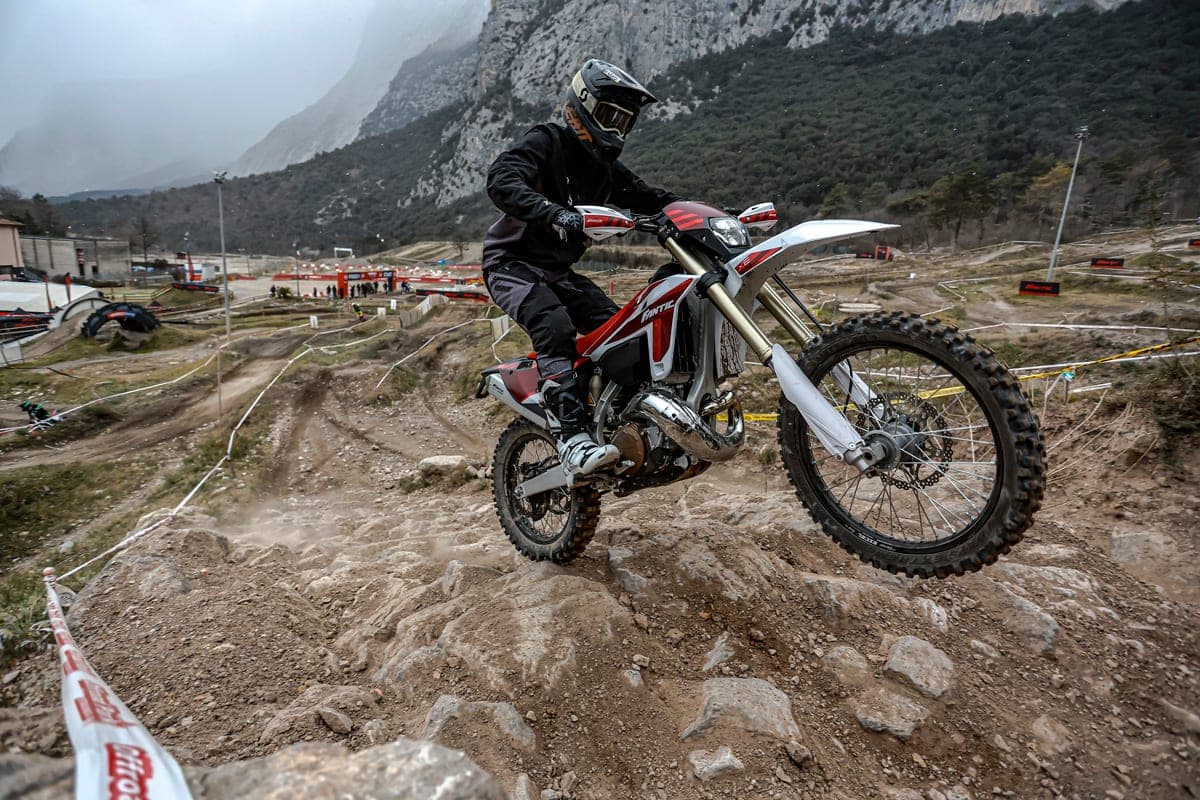 Rider in black gear popping the front wheel over rocks on a 2027 Fantic XE 300, mountain terrain and race tape visible behind