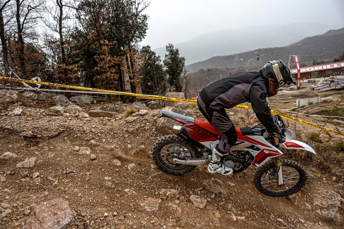 Fantic XE 300 leaning downhill through a rocky, tape-lined enduro section, rider crouched low with the rear shock compressed