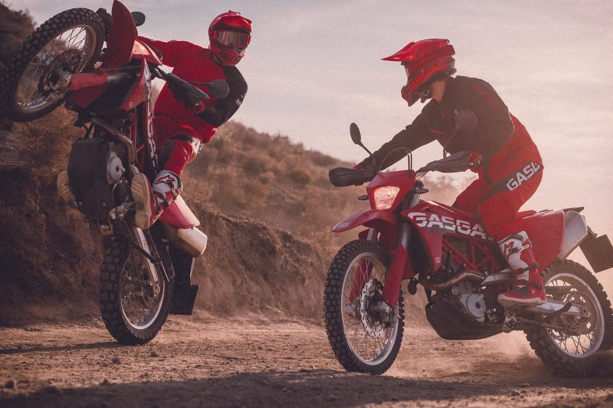 Two riders in red and black GasGas gear popping wheelies on a dusty hillside track, one lifting the front wheel high off the ground