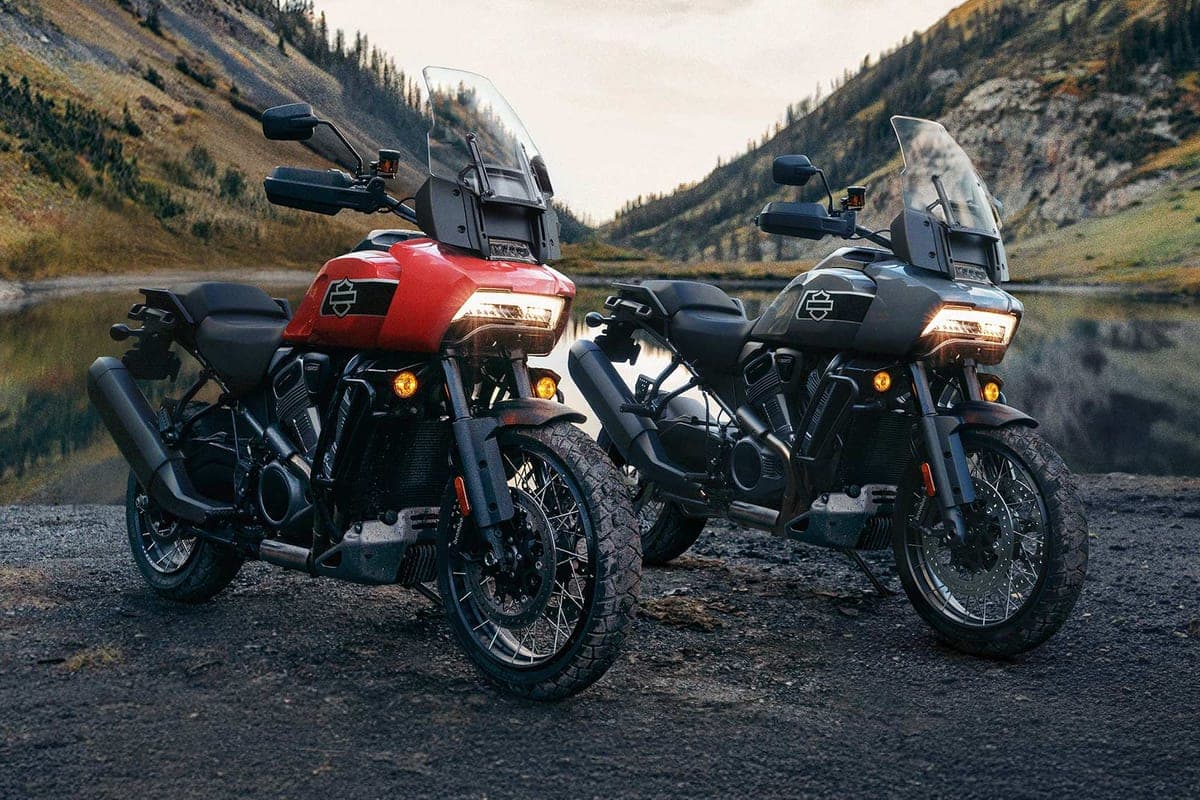 Two Harley-Davidson Pan America 1250 Specials, one orange and one grey, parked front three-quarter beside a mountain lake with headlights on