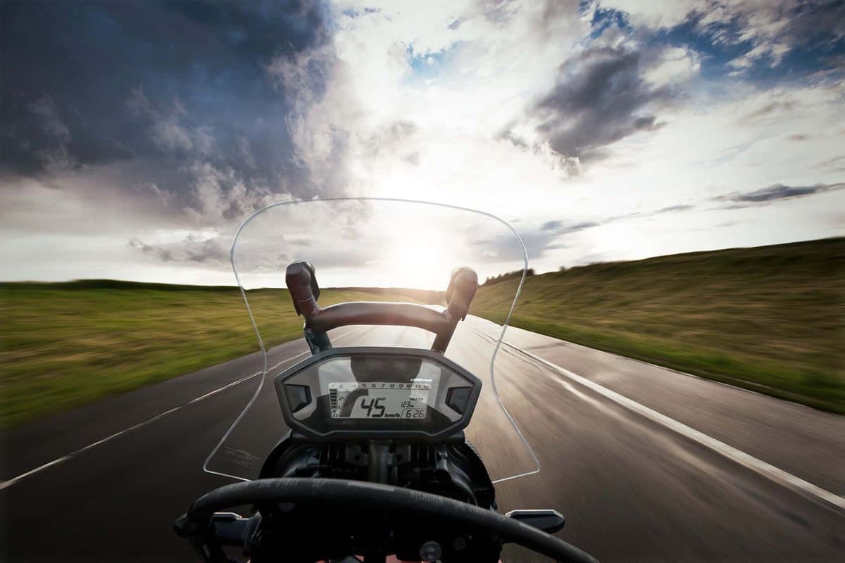 First-person cockpit view speeding down a sunlit rural highway on a Honda CRF250L Rally, showing the digital gauge cluster and windscreen