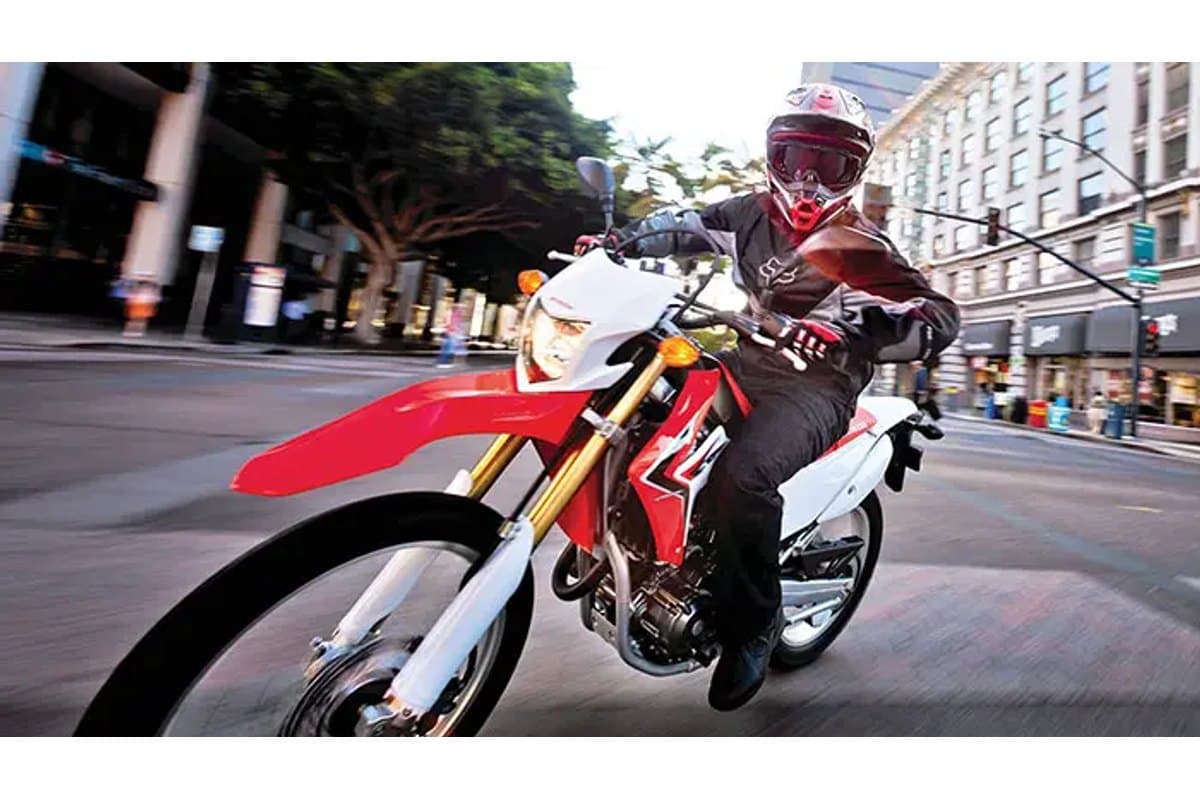 Rider in Fox riding gear leaning into a city street turn on a red and white Honda CRF250L, front wheel lifting slightly off the pavement