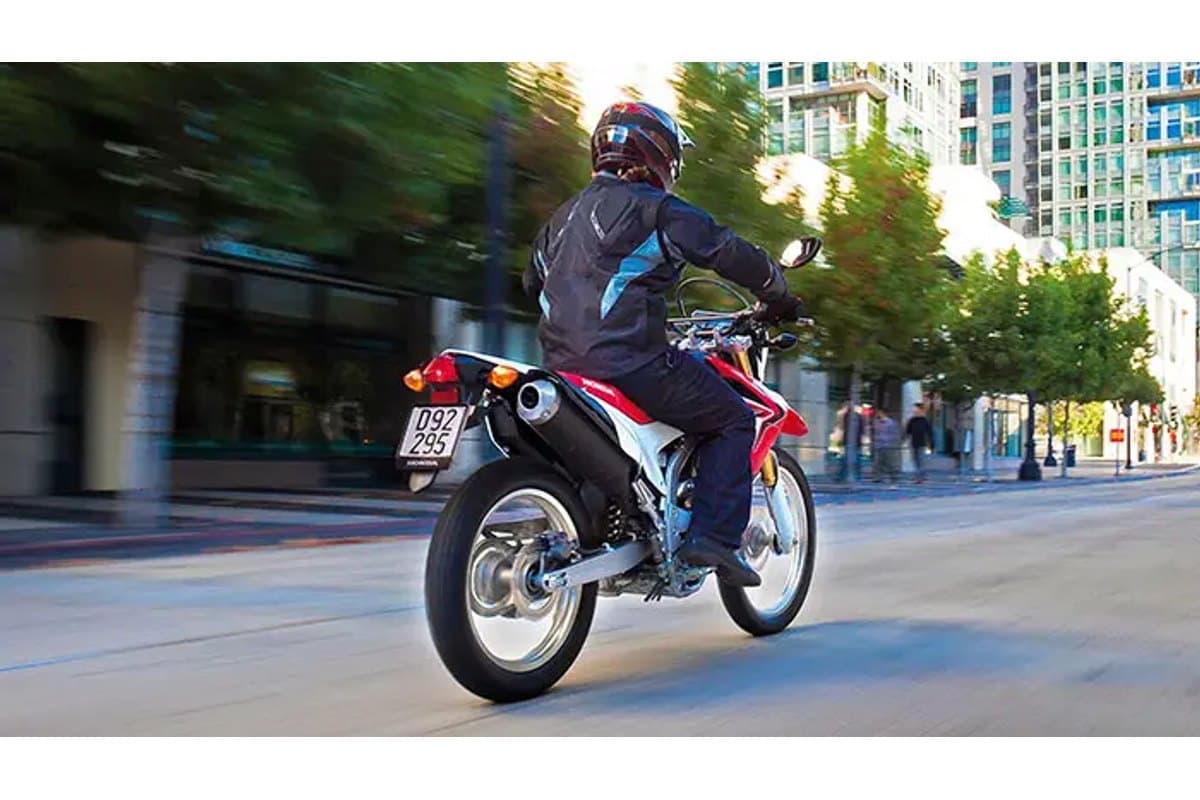 Rear three-quarter view of a Honda CRF250L rounding a city corner, rider in dark jacket with reflective panels, exhaust and taillight visible