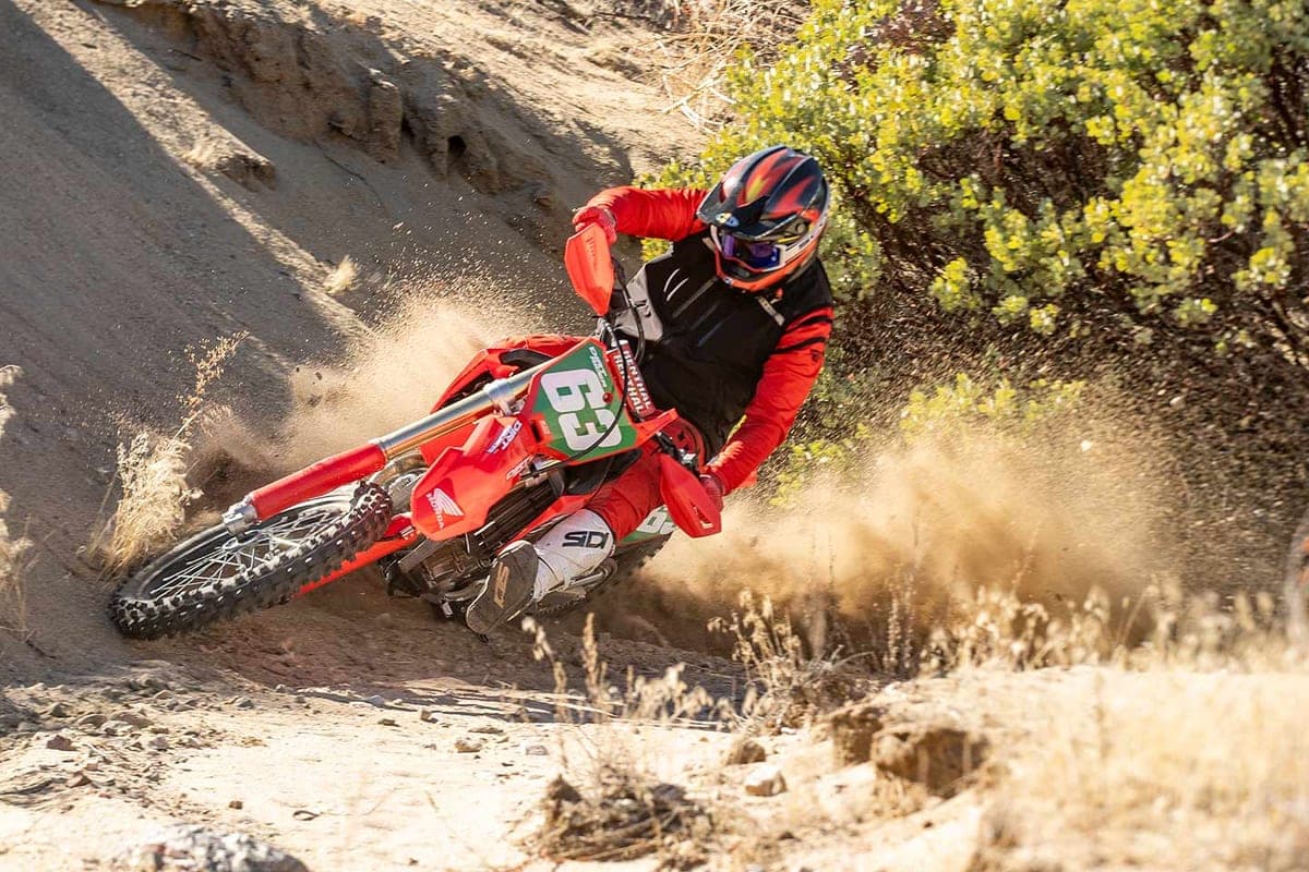 Rider in red and black gear leaning a Honda CRF250RX through a dusty desert turn, roosting sand off the rear knobby tire
