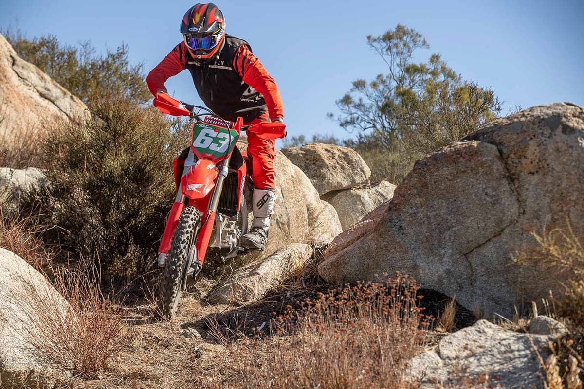 Honda CRF250RX climbing a rocky, boulder-strewn trail with the rider standing on the pegs under a clear blue sky